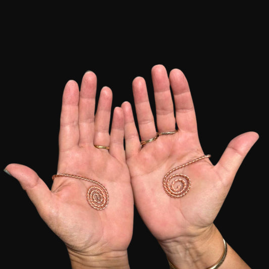 Two hands displaying copper spiral EMF hand healing cuffs against a black background