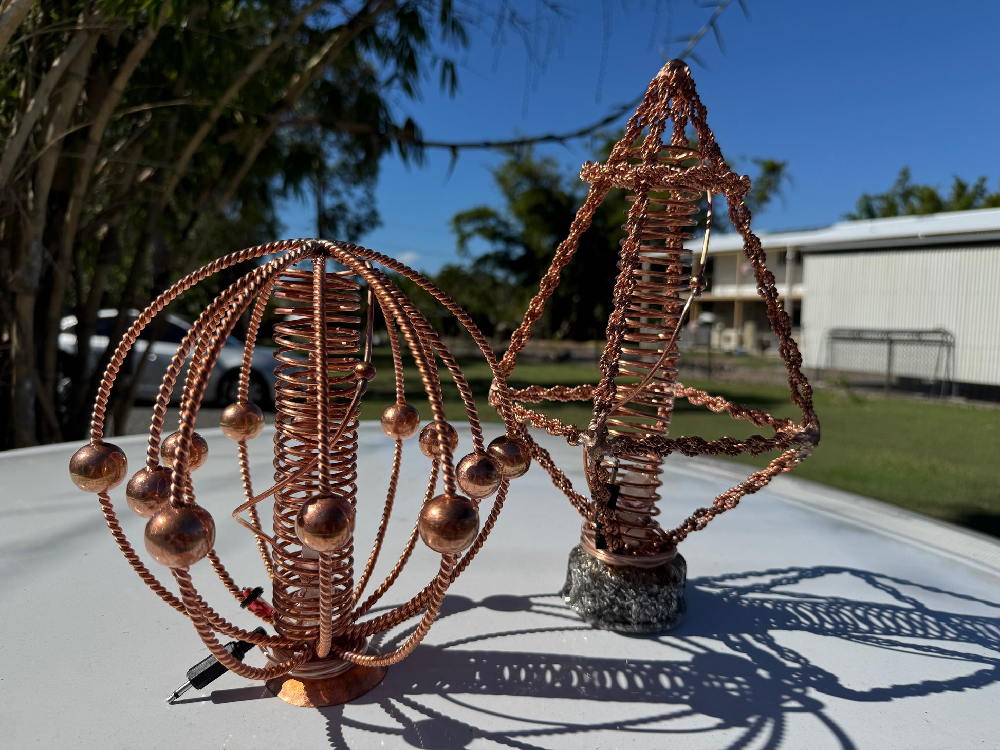 Copper wire sculpture on a table outdoors with trees and building in the background